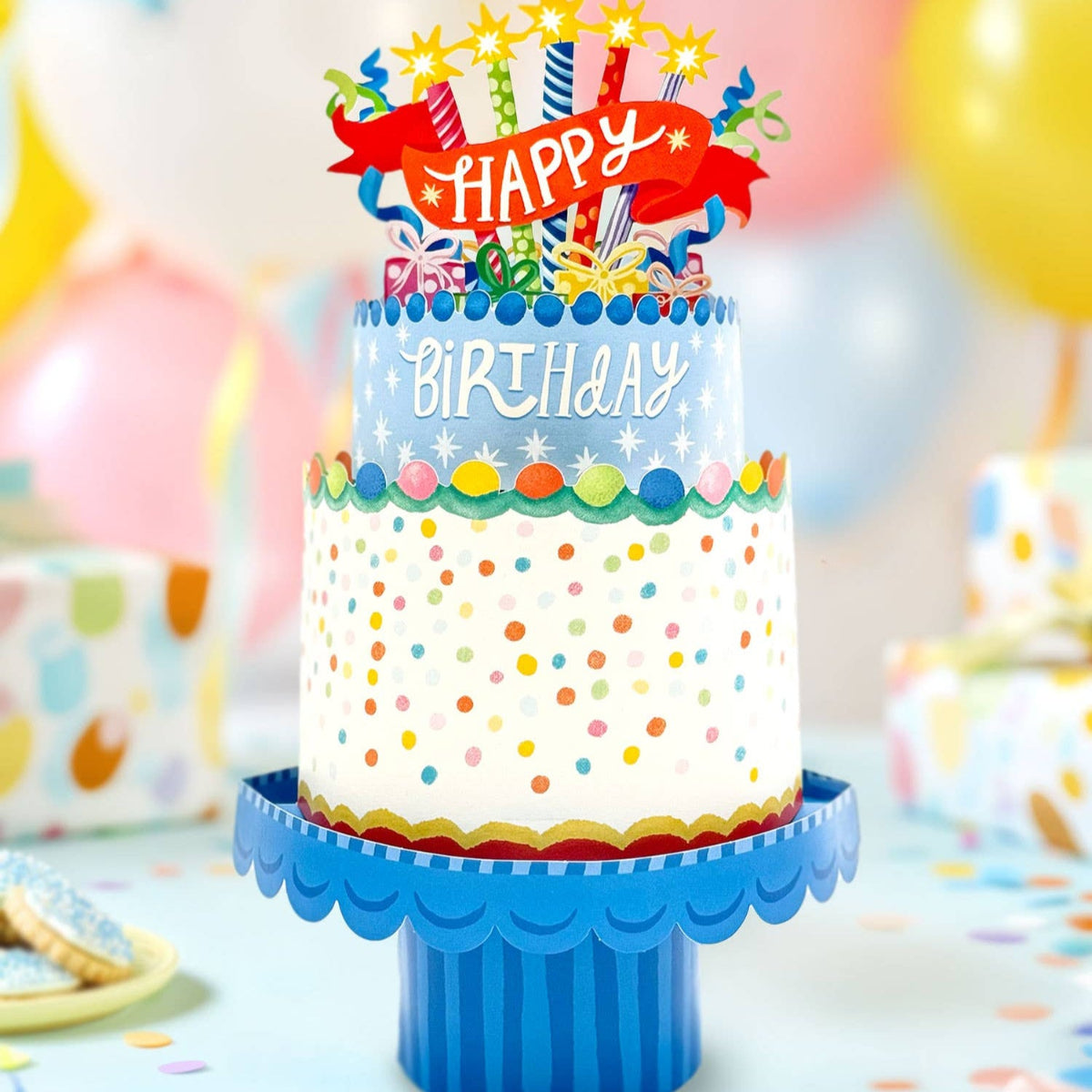 Colorful birthday cake with 'Happy Birthday' topper on a blue stand, surrounded by balloons and party decorations.