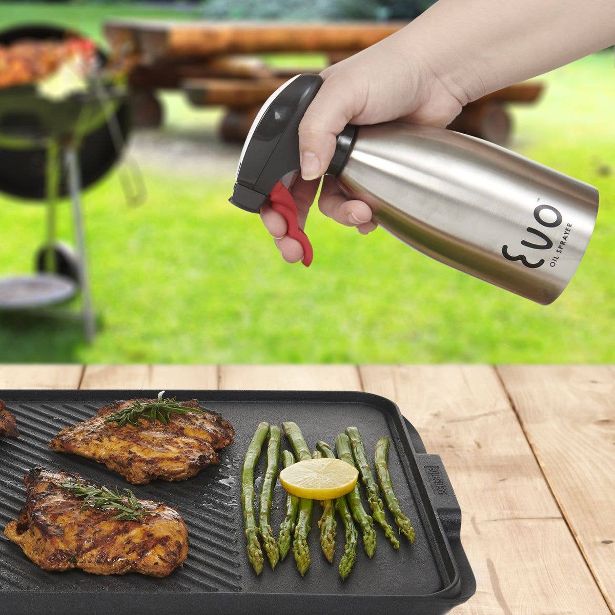 Person using a stainless steel oil sprayer over grilled vegetables and meats on a grill pan.