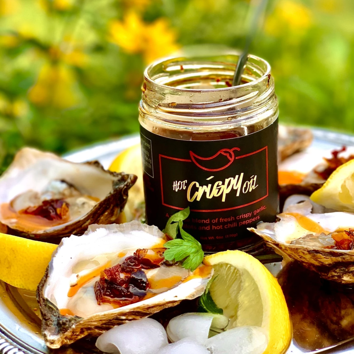 Jar of oyster sauce on a plate with oysters and lemon wedges, outdoor setting with yellow flowers.