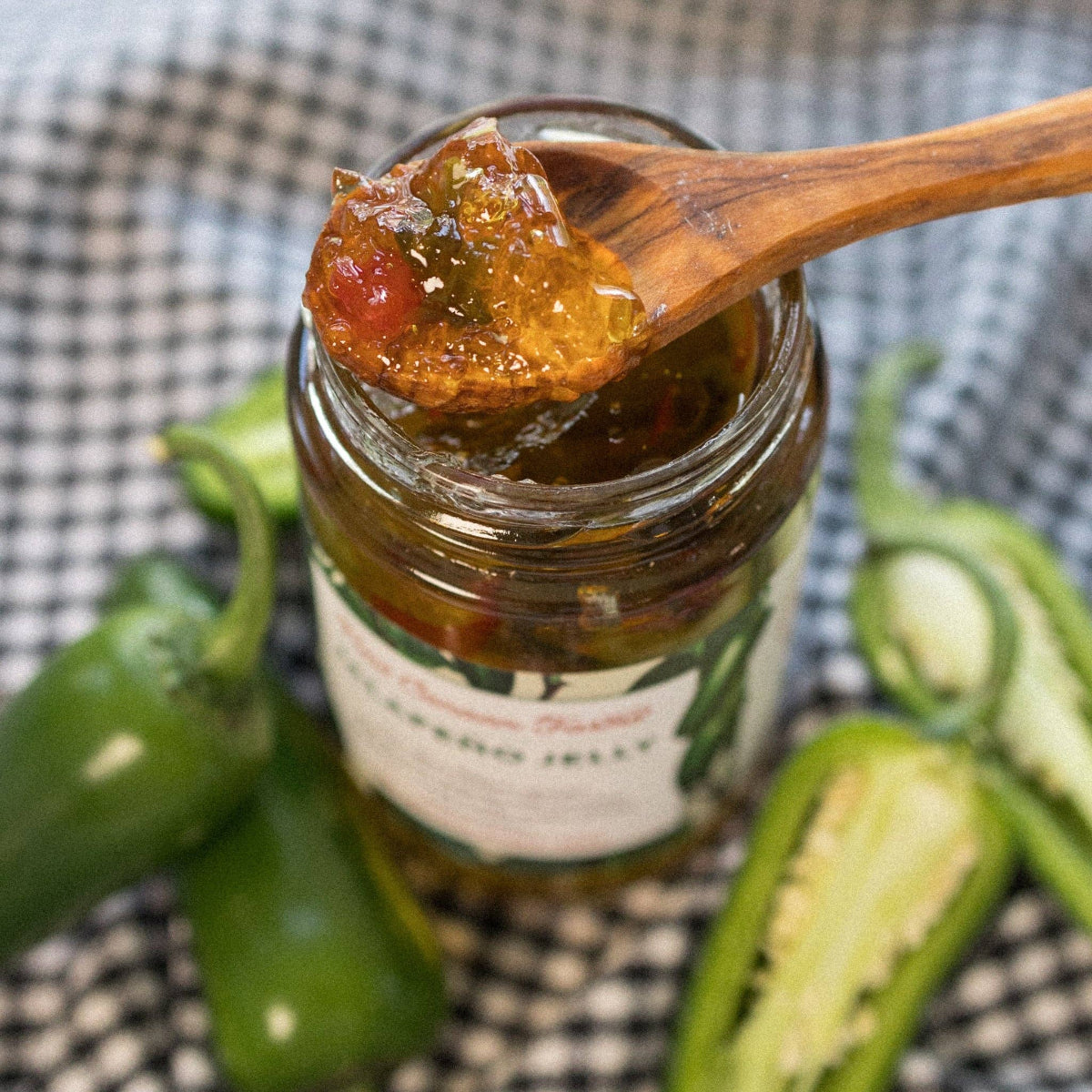 Jar of jalapeño jelly with a wooden spoon, surrounded by jalapeños on a checkered background