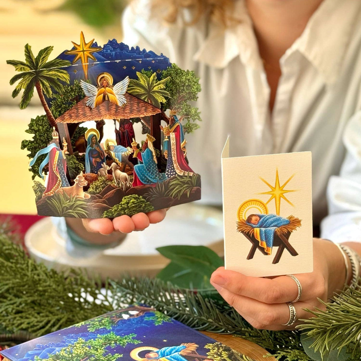 Person holding a 3D nativity scene card and a traditional nativity scene card with a festive background.