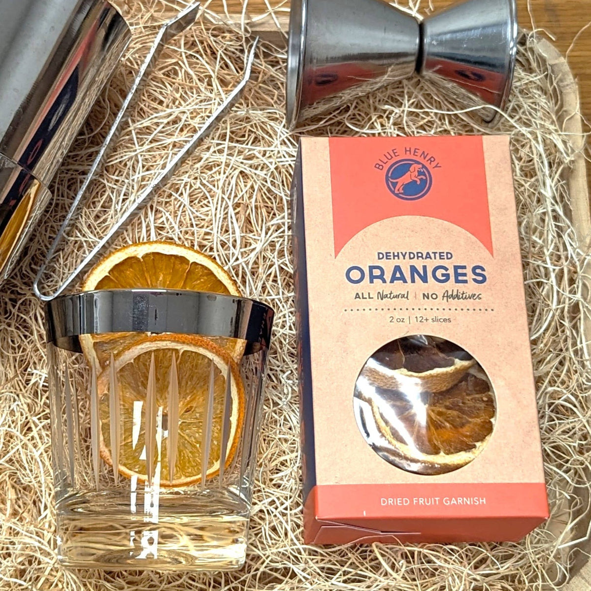 Cocktail shaker, glass with orange slices, and box of dehydrated oranges on a wooden surface.
