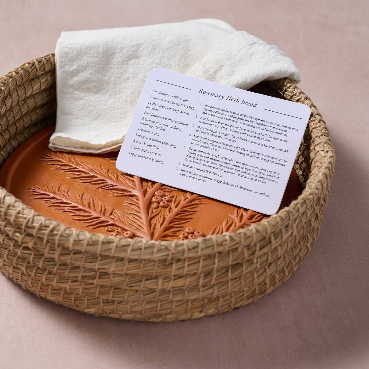 A white recipe card titled 'Rosemary Herb Bread' rests on a terracotta-colored clay loaf in a woven straw basket, next to a folded white cloth on a soft beige background.