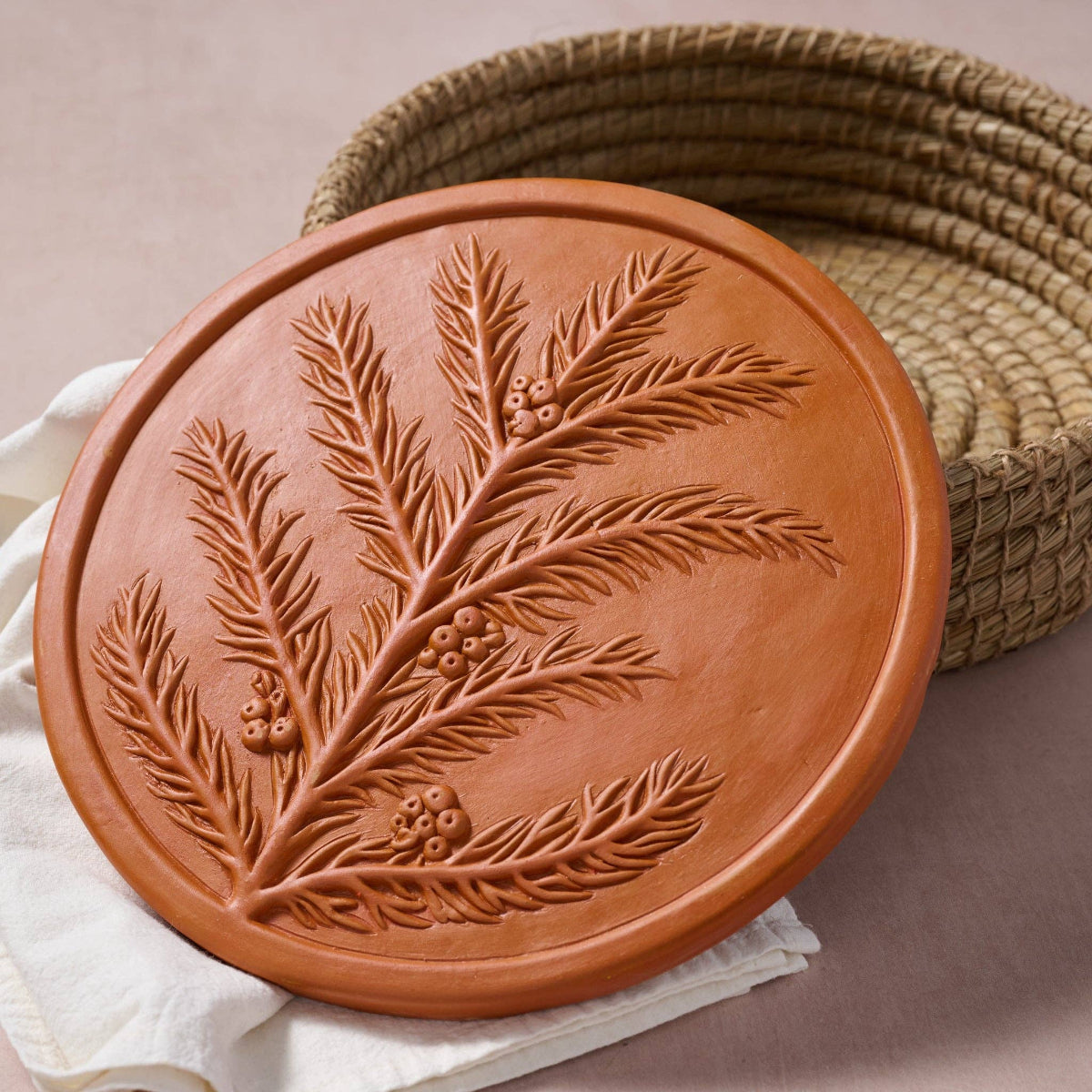 Round terracotta decorative plate with embossed evergreen branches and berries, styled on a white cloth next to a woven basket.