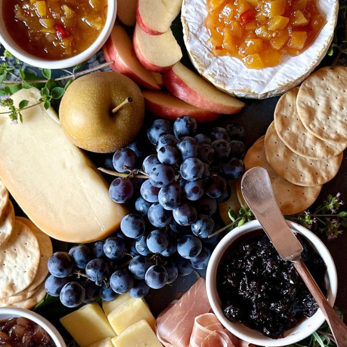 Platter with assorted cheeses, fruits, and chutneys on a wooden board with 'Tait Farm Cheese Plavings' text.