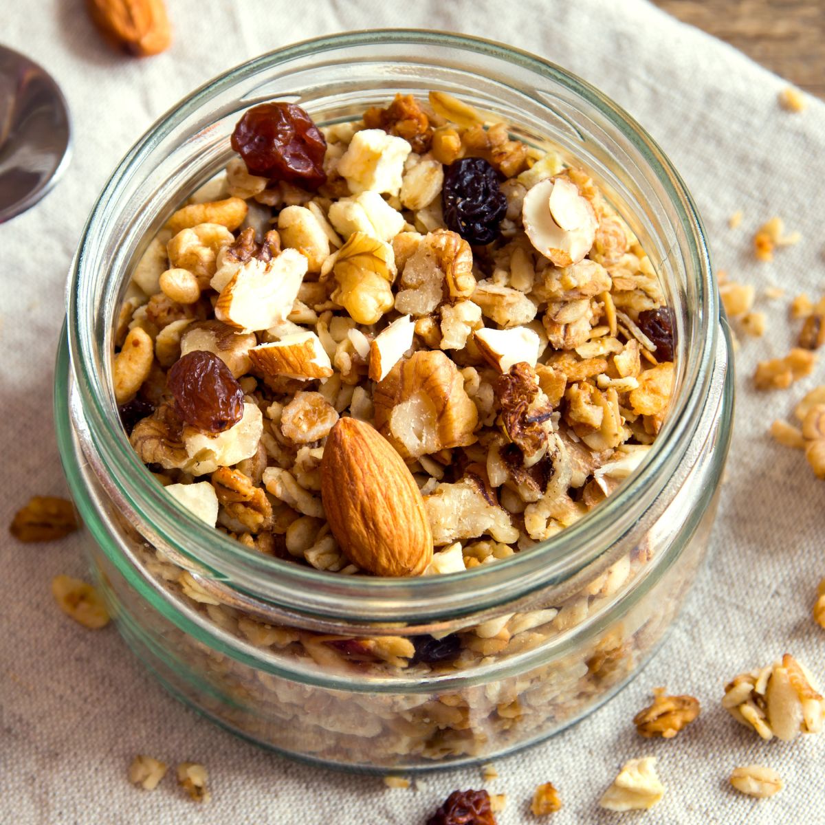 Glass jar filled with granola, nuts, and raisins on a light fabric background