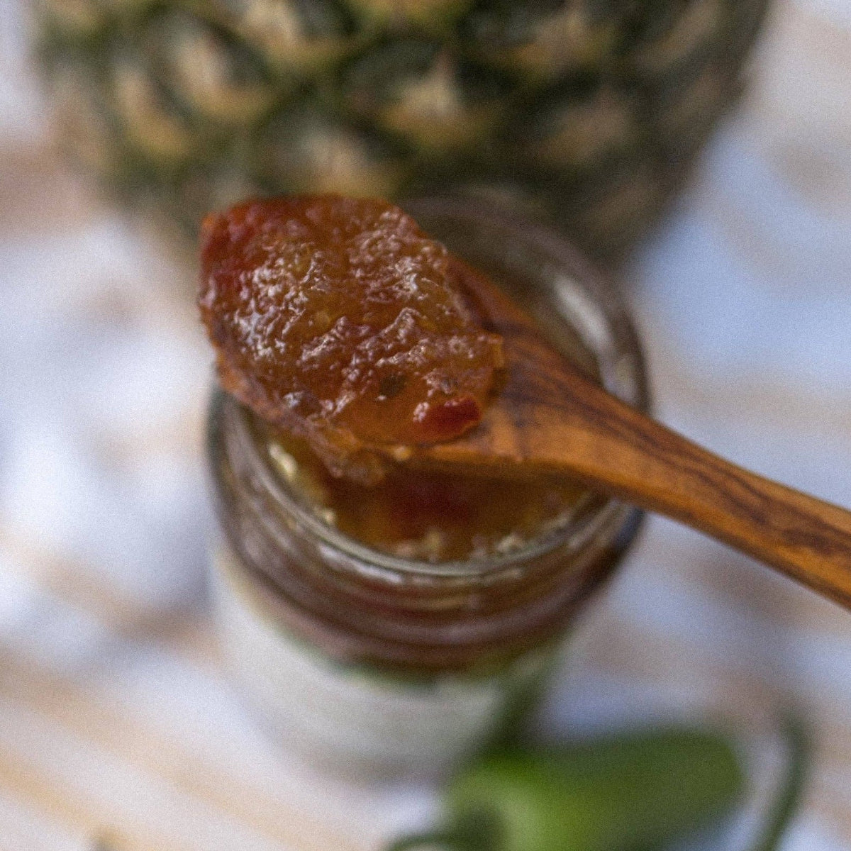 Wooden spoon with jam over a jar, pineapple and jalapeño in the background