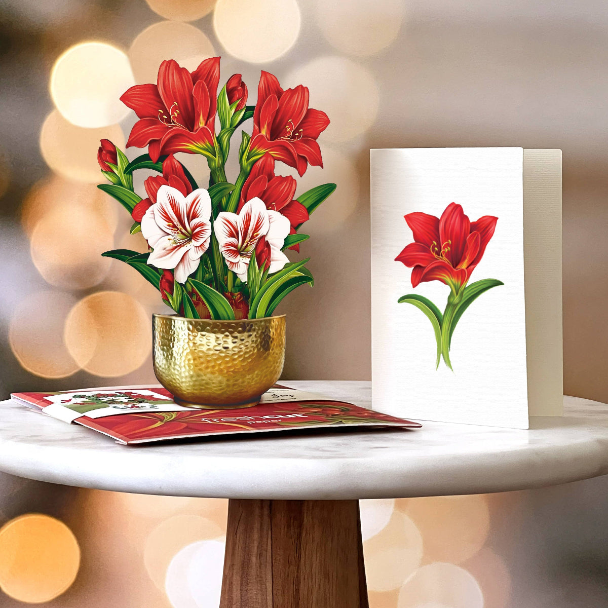 Decorative setup with a gold pot of red and white flowers and matching cards on a blurred light background.