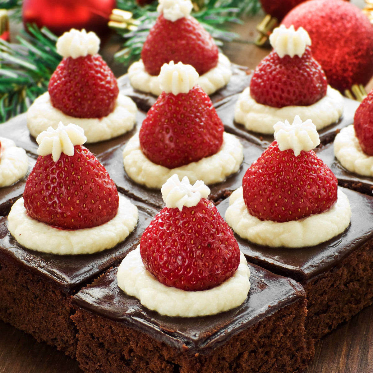 Chocolate brownies topped with strawberries and cream, decorated like Santa hats, on a wooden surface.