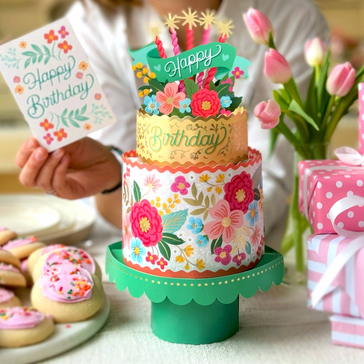 Woman holding a birthday card next to a decorated birthday cake with flowers and candles.