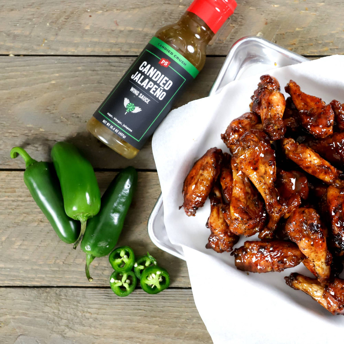 Bottle of candied jalapeño wing sauce with wings and jalapeños on a wooden surface