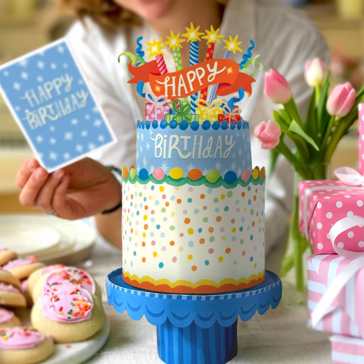 Colorful birthday cake with candles and a 'Happy Birthday' card held by a person.