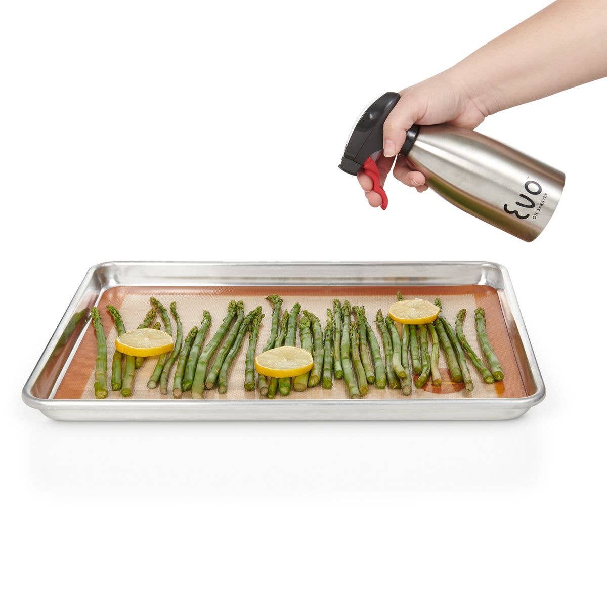 Person using a cooking spray bottle over a tray of asparagus with lemon slices on a white background