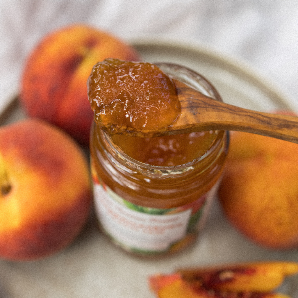 Jar of peach jam with a wooden spoon, surrounded by peaches on a white plate.