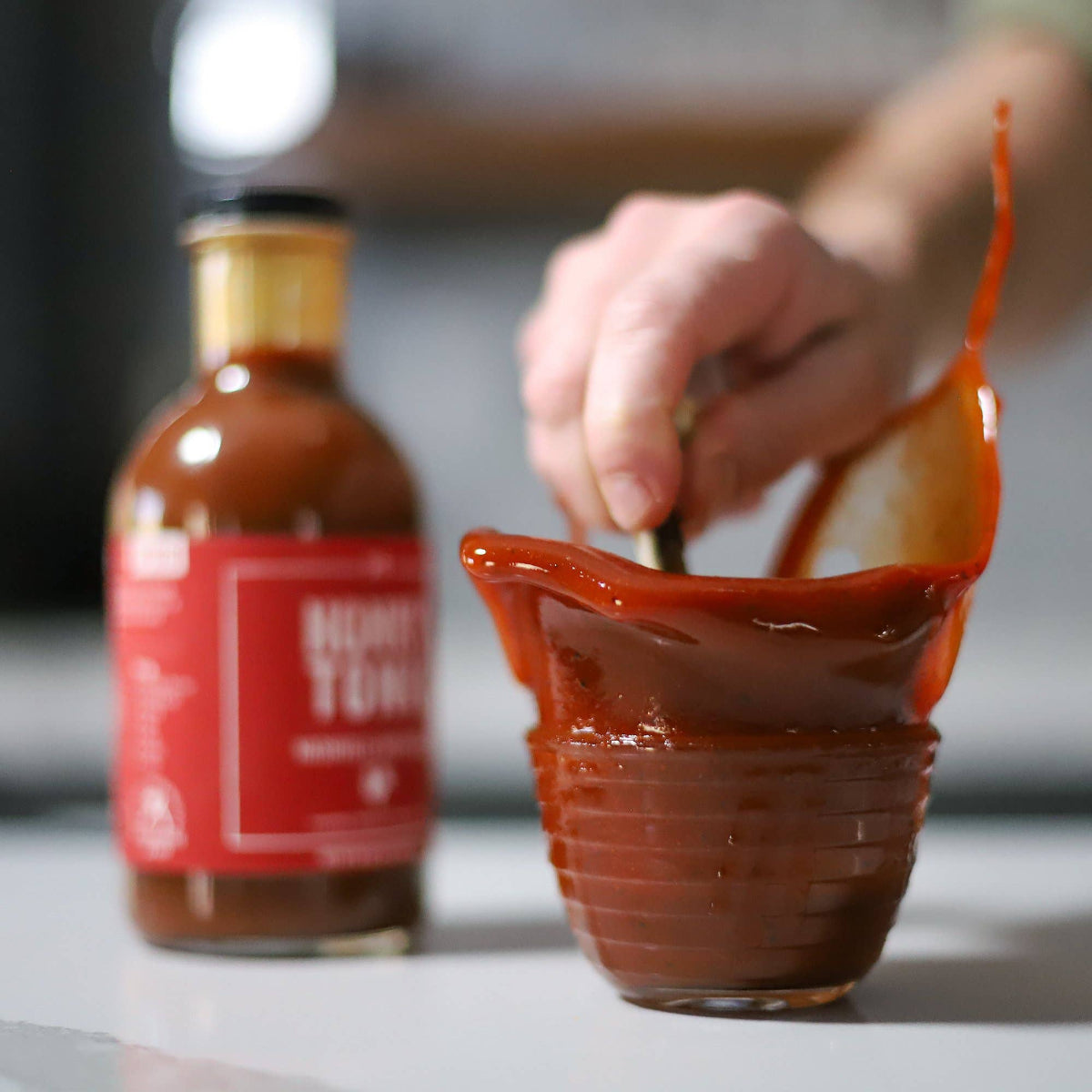 Person using a bottle to pour hot sauce into a container with a bottle of hot sauce labeled 'HOT' in the background.