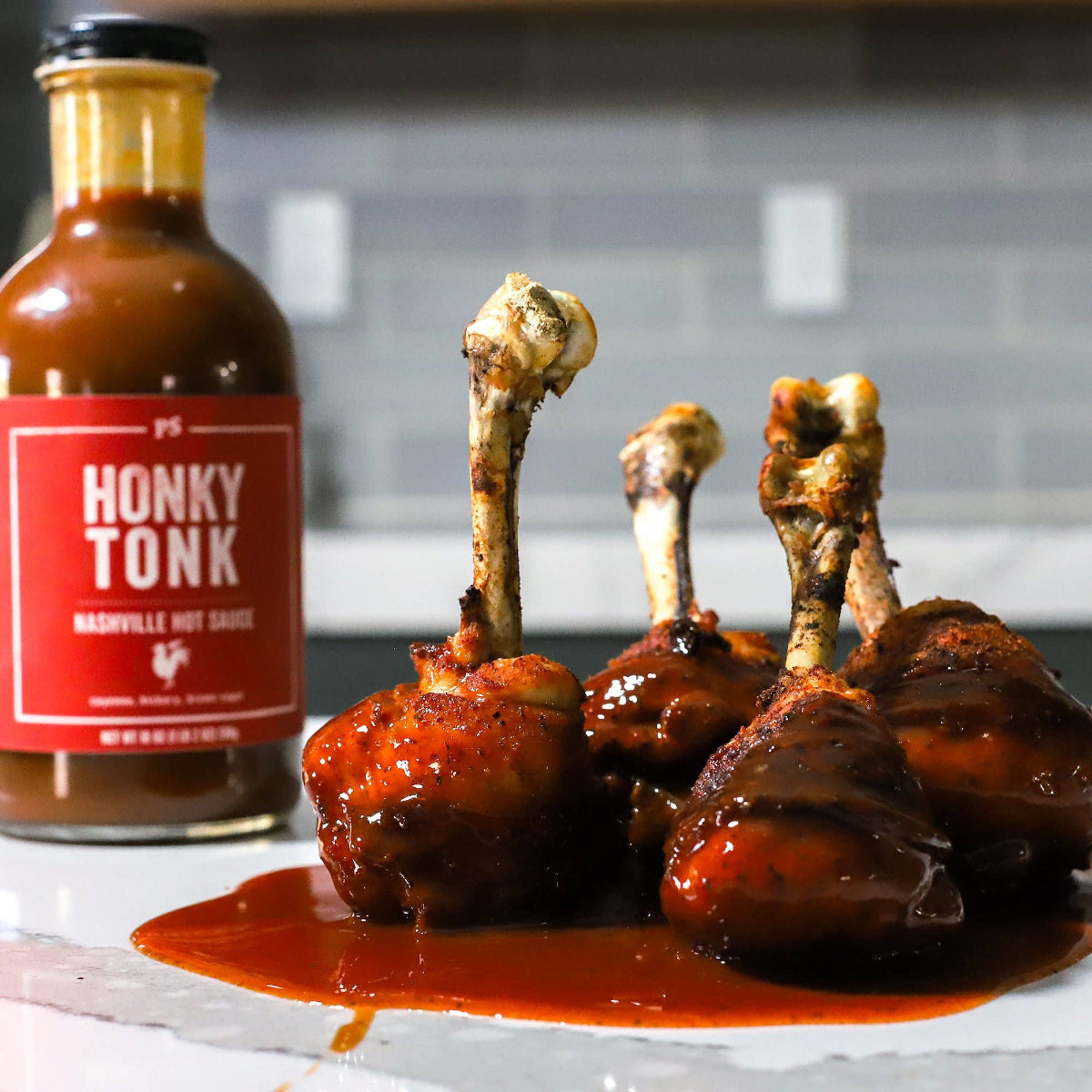 Braised chicken drumsticks with a bottle of 'Honky Tonk' hot sauce on a marble surface.