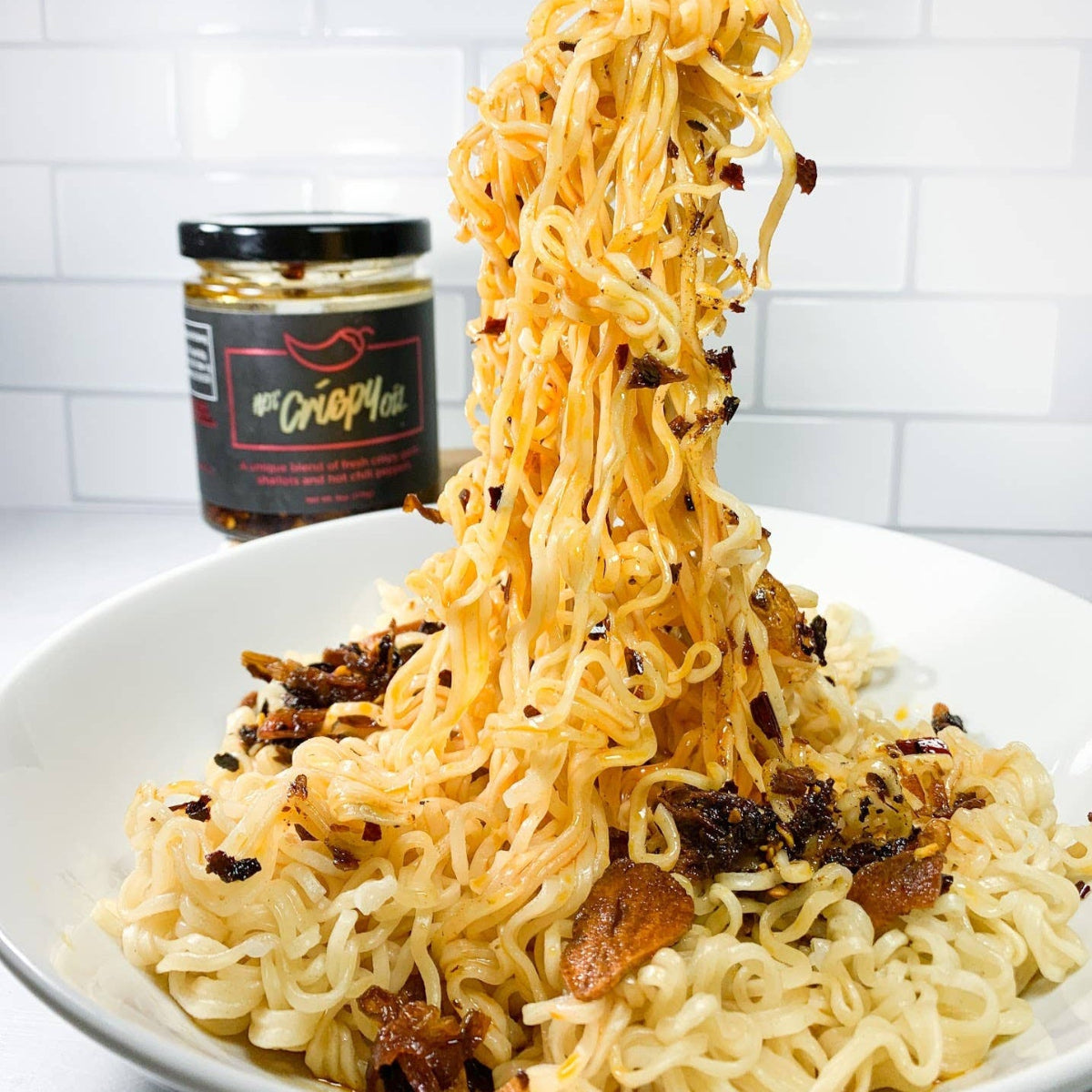 Bowl of noodles with a jar of sauce in the background on a white tiled wall.