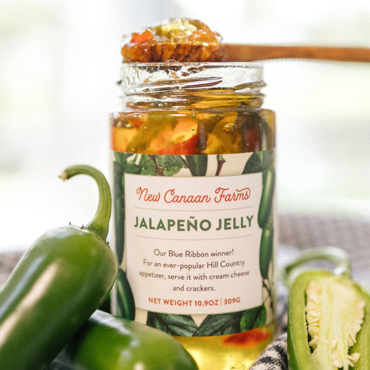 Jar of New Canaan Farms Jalapeño Jelly with a spoonful of jelly and jalapeño peppers around it.