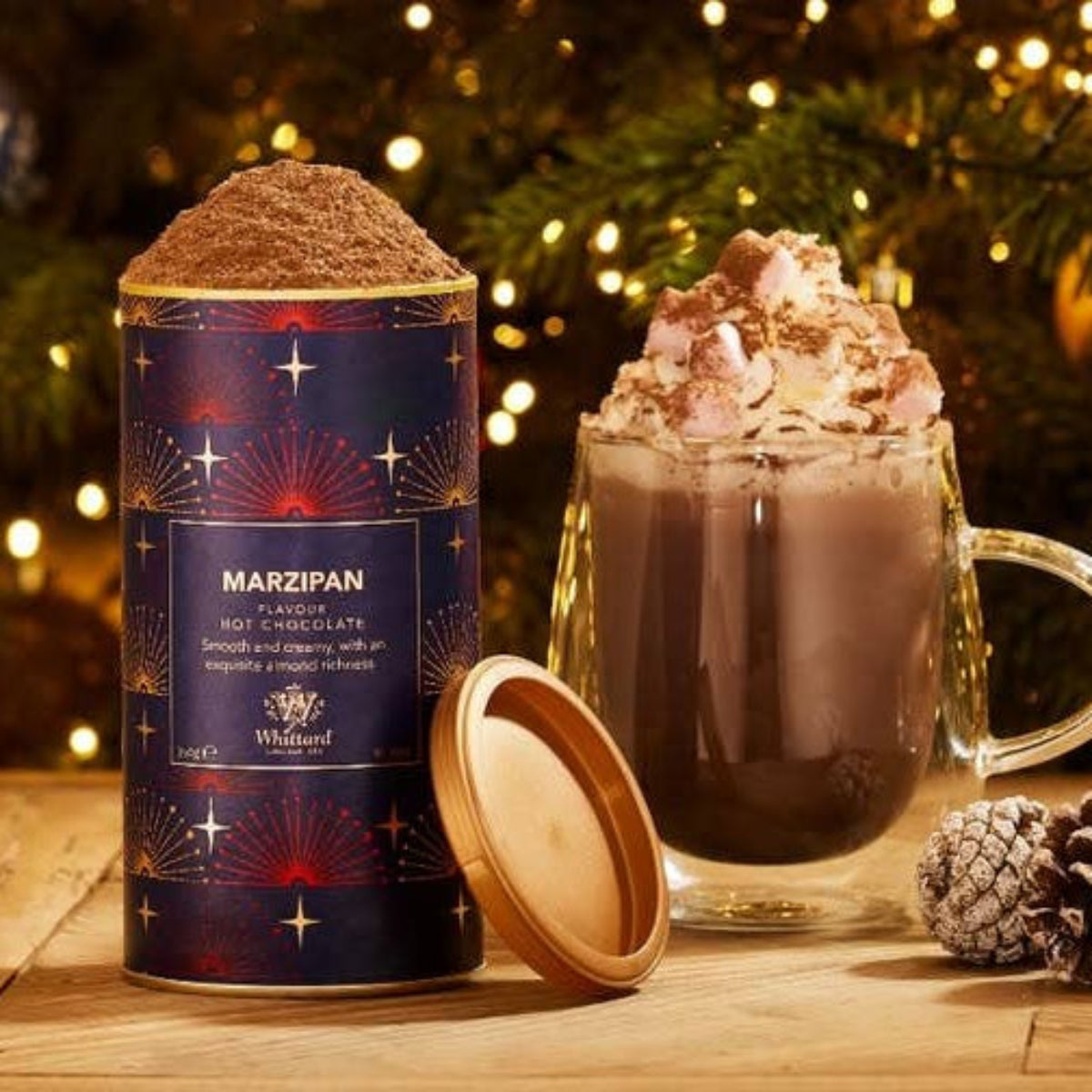 Hot chocolate with whipped cream and a can of Marzipan hot chocolate mix in front of a Christmas tree.