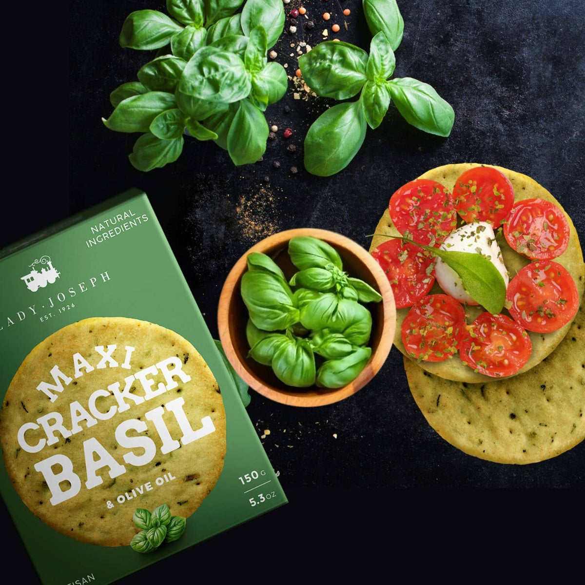Box of Lady Joseph Maxi Cracker Basil with basil leaves and a cracker topped with tomatoes and cheese on a dark surface.