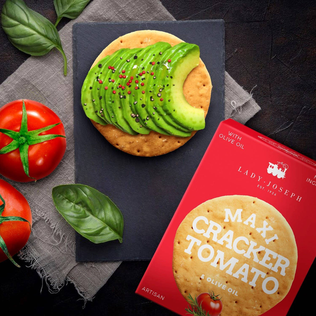 Maxi cracker with avocado and tomatoes on a dark surface, next to a box of Lady Joseph Maxi Cracker Tomato.
