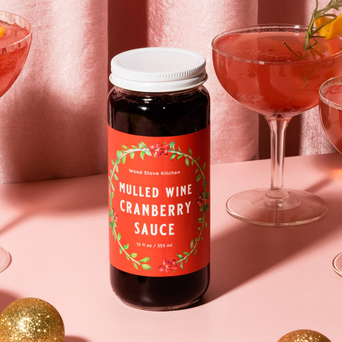 Mulled wine cranberry sauce bottle with cocktail glasses and festive ornaments on a pink background