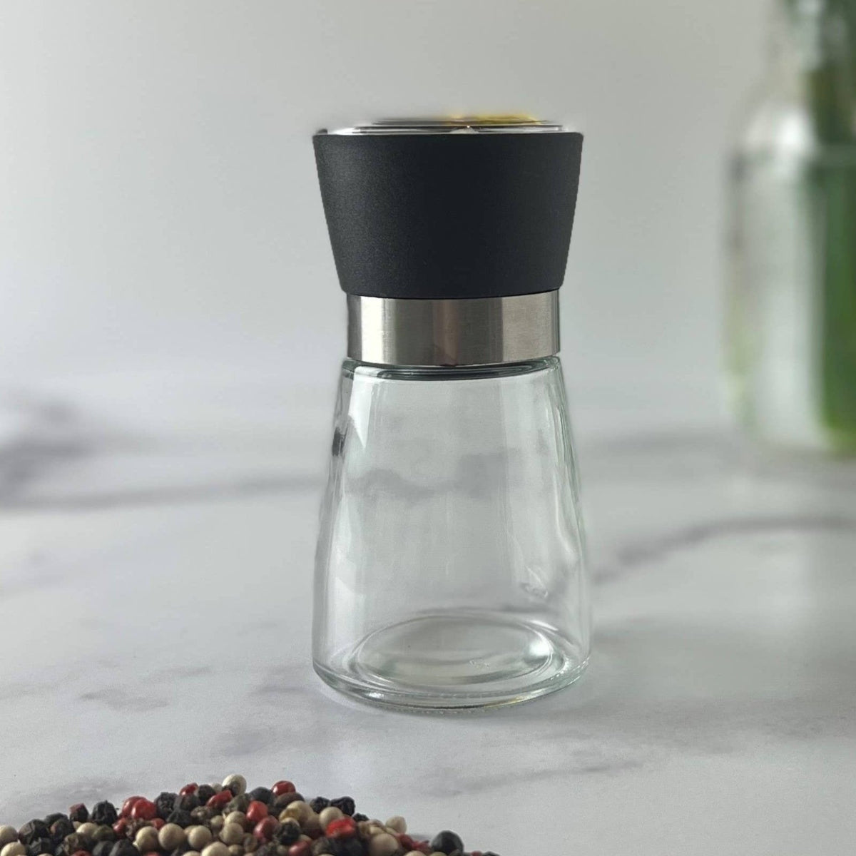 Clear pepper grinder with black top on a marble surface with yellow flowers in the background