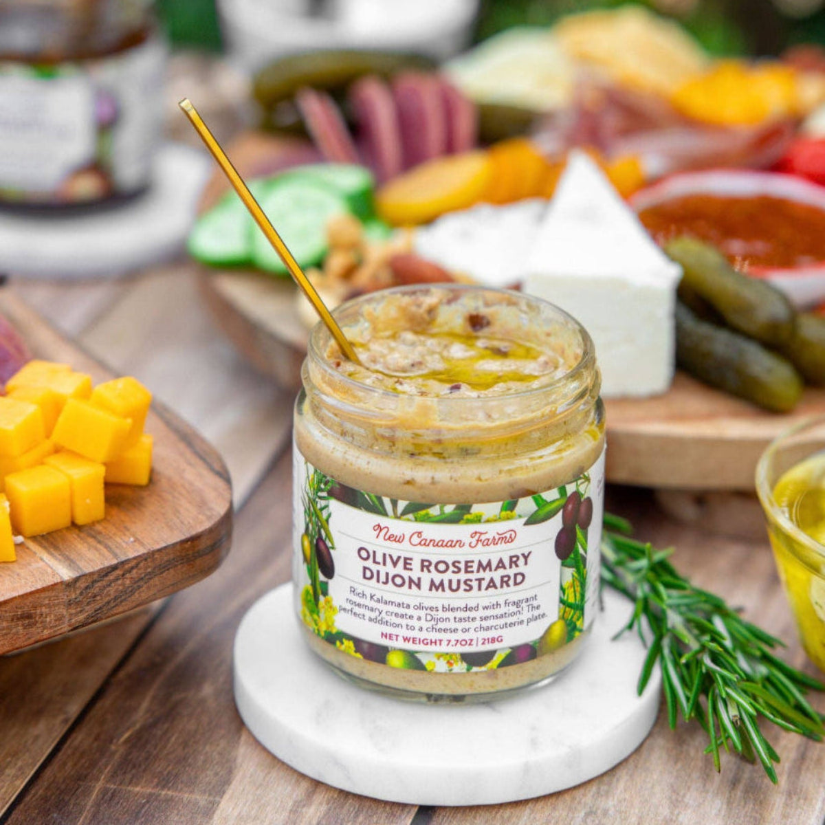 Jar of olive rosemary dijon mustard on a wooden table with a platter of assorted foods.