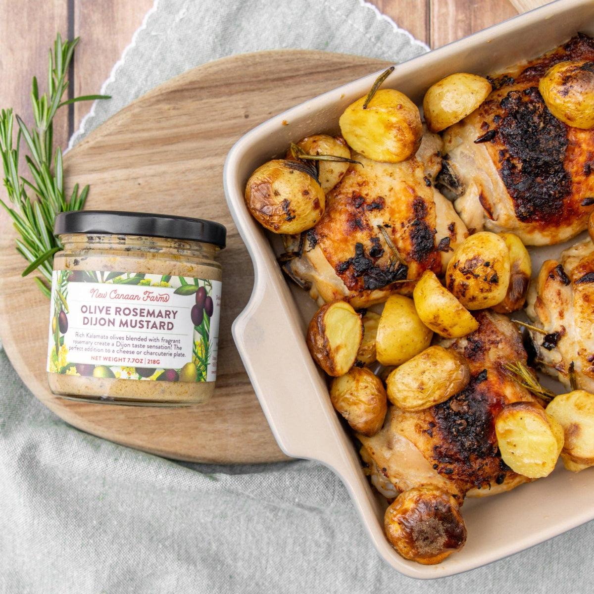 Baked chicken and potatoes with a jar of olive rosemary dijon mustard on a wooden board.