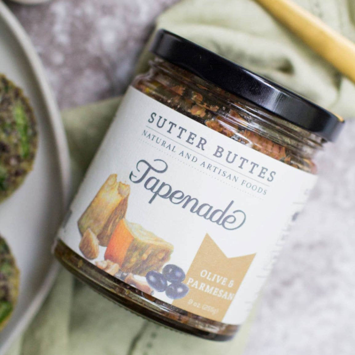 Jar of Sutter Buttes Tapenade on a textured surface with a spoon and dish in the background.