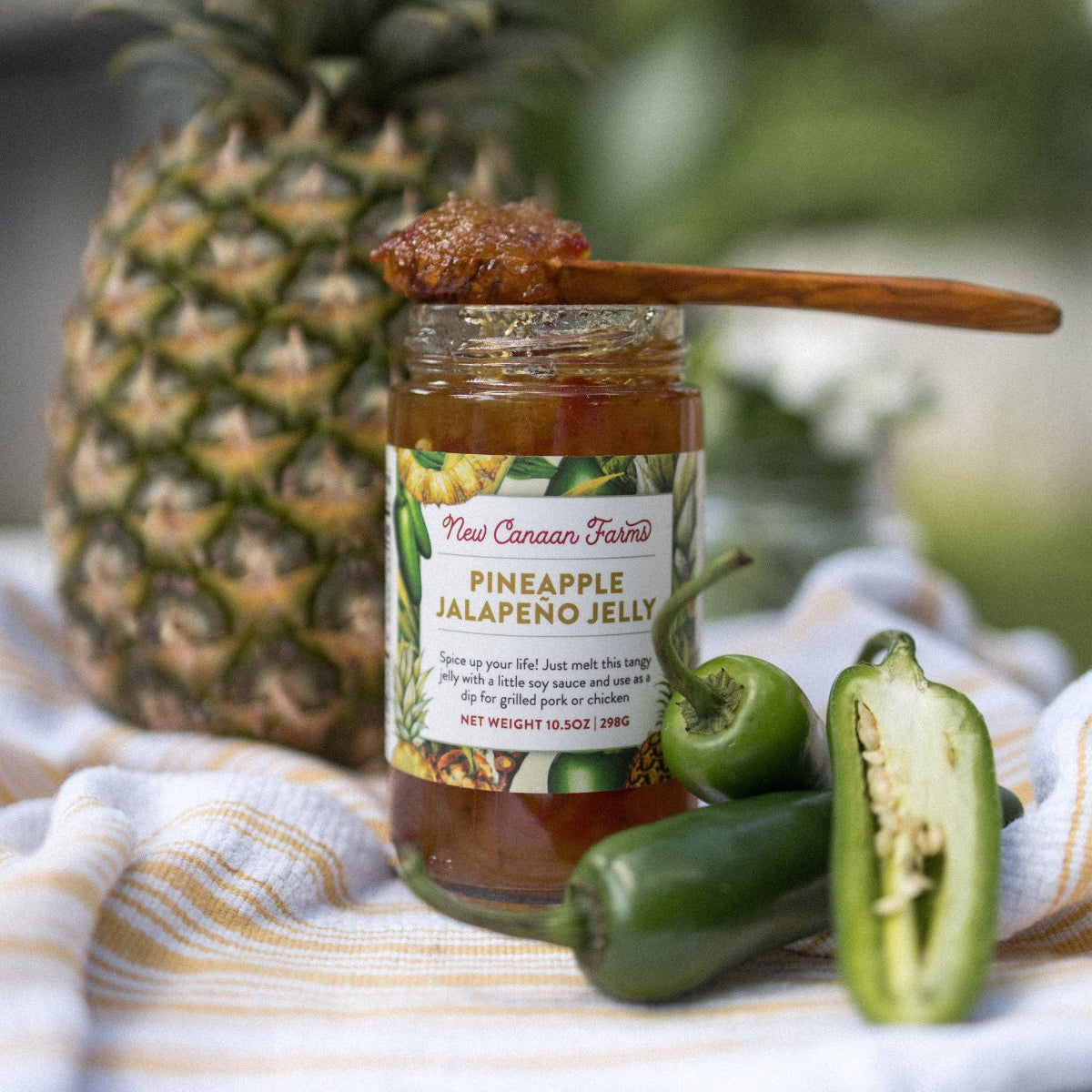 Jar of Pineapple Jalapeno Jelly with a wooden spoon, surrounded by jalapenos and a pineapple on a striped cloth.