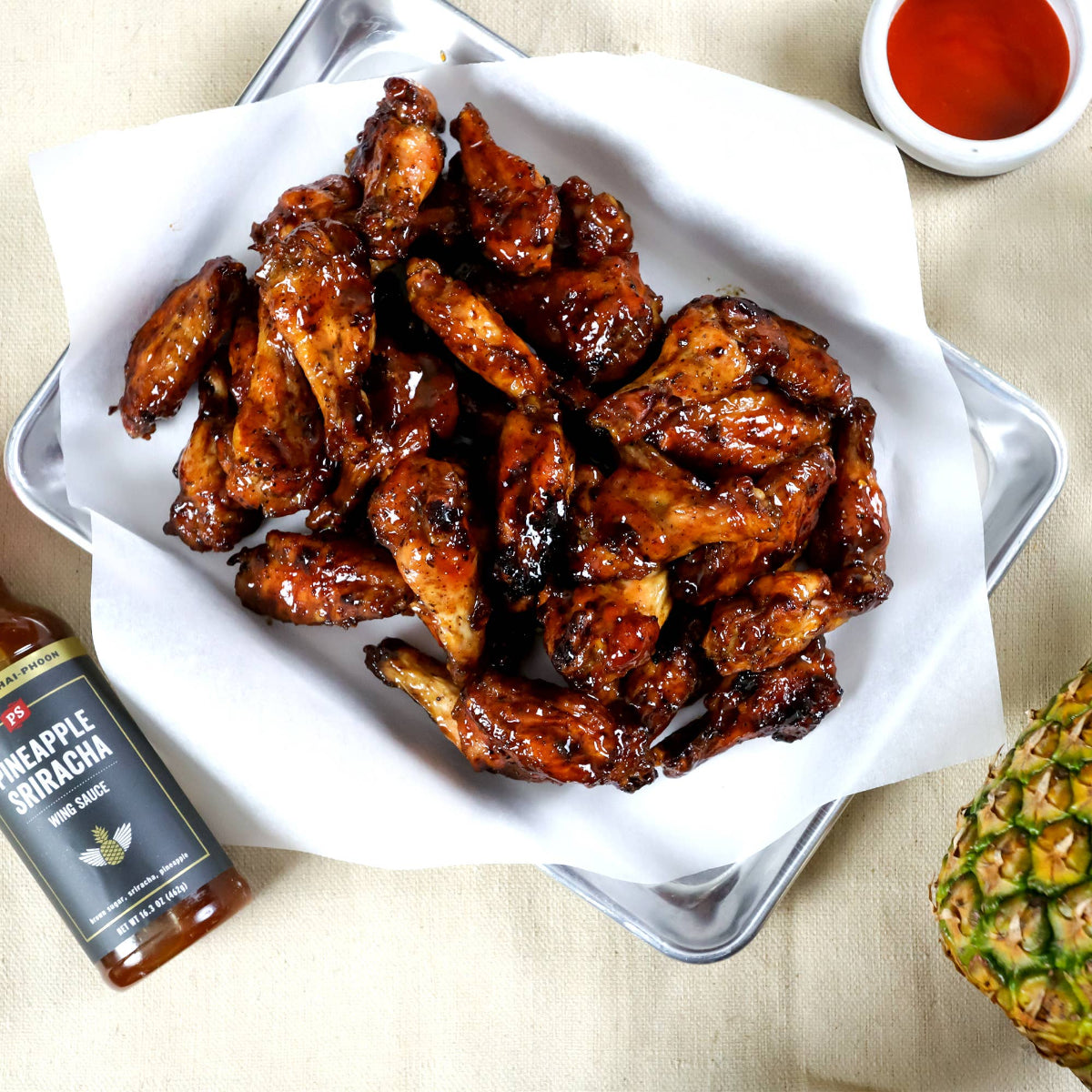 Baked chicken wings with pineapple sriracha sauce on a white plate, accompanied by a bottle of sauce and a pineapple wedge.