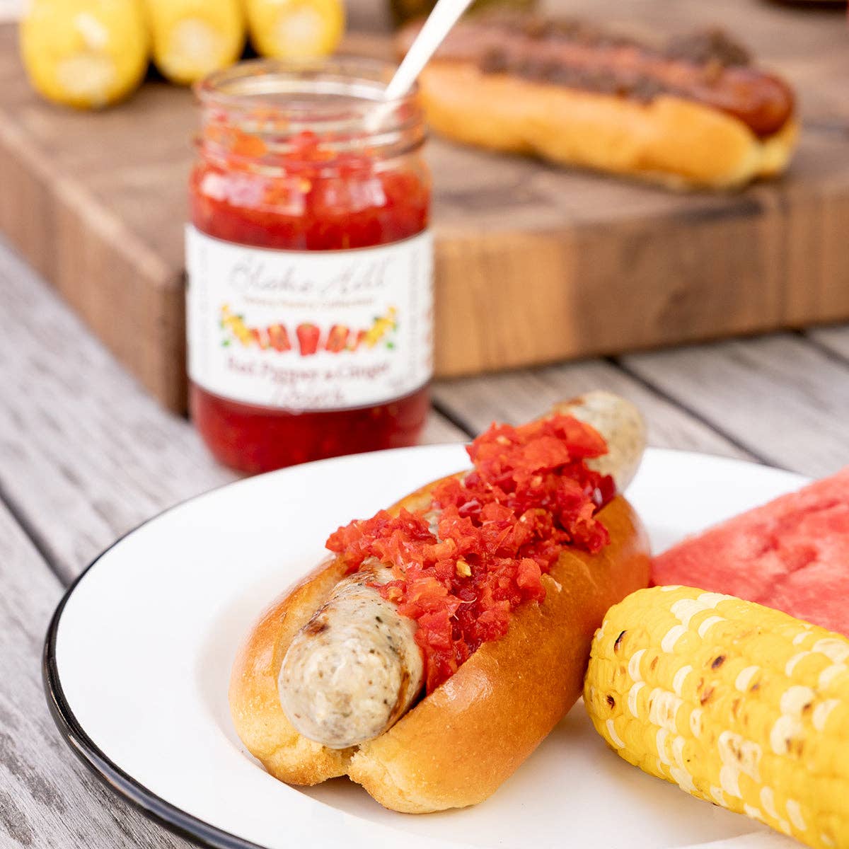 Hot dog with relish on a bun on a plate, corn on the cob, and a jar of relish in the background.