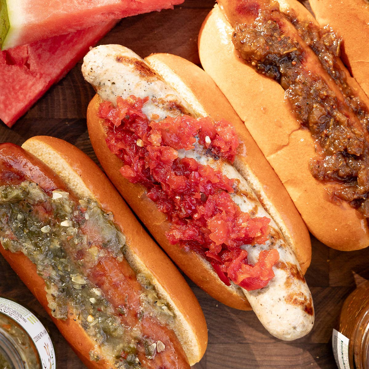 Hot dog with relish on a bun on a plate, corn on the cob, and a jar of relish in the background.