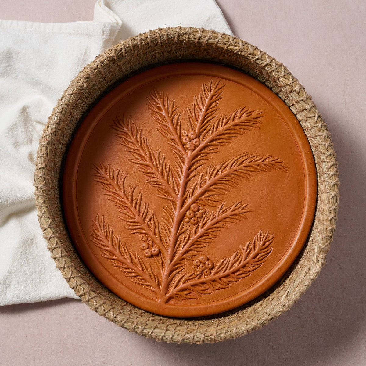 A round terracotta clay plate with an embossed evergreen branch design, nested inside a woven straw basket, with a white cloth in the background.