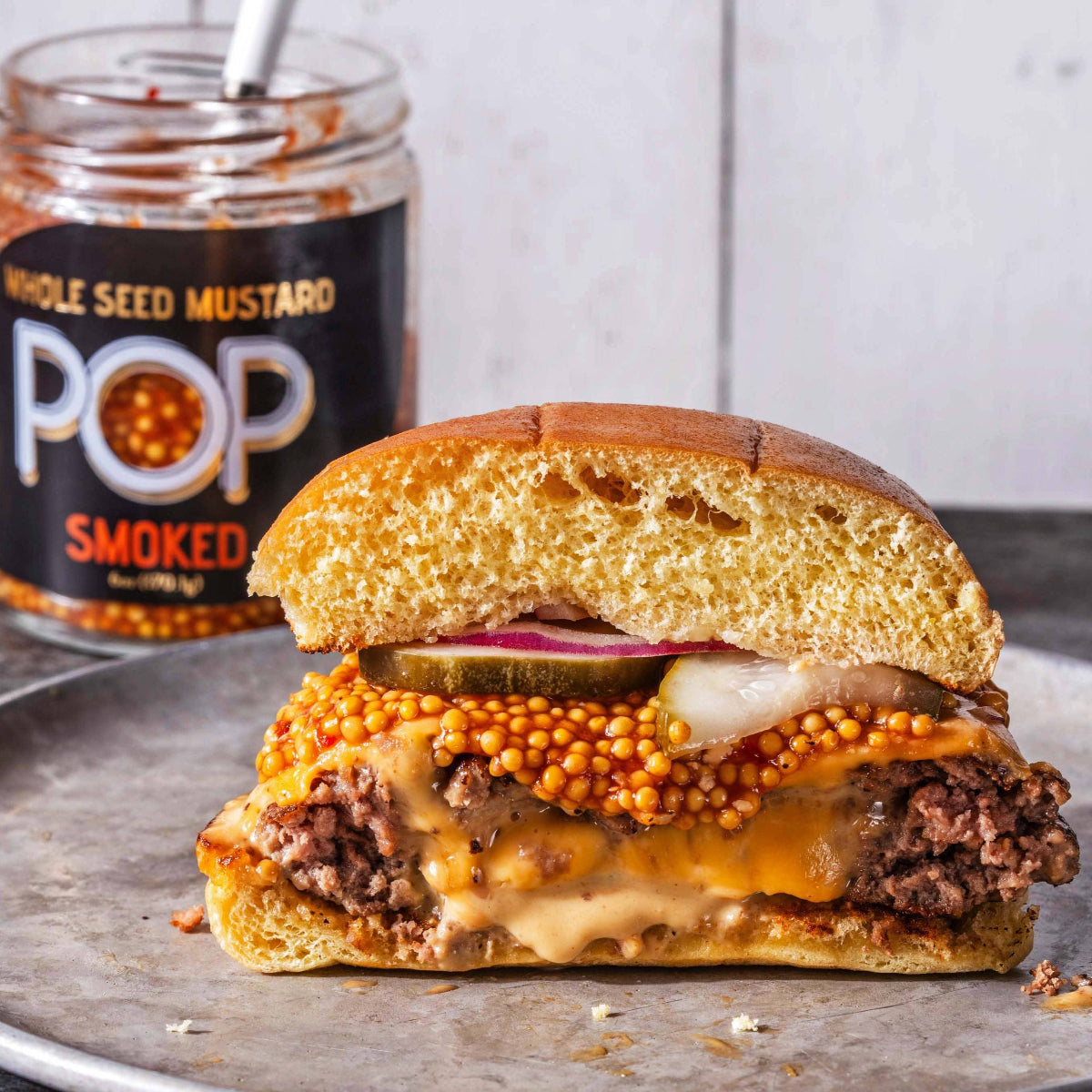 Hamburger cut in half on a plate with a jar of 'POP Smoked Whole Seed Mustard' in the background.