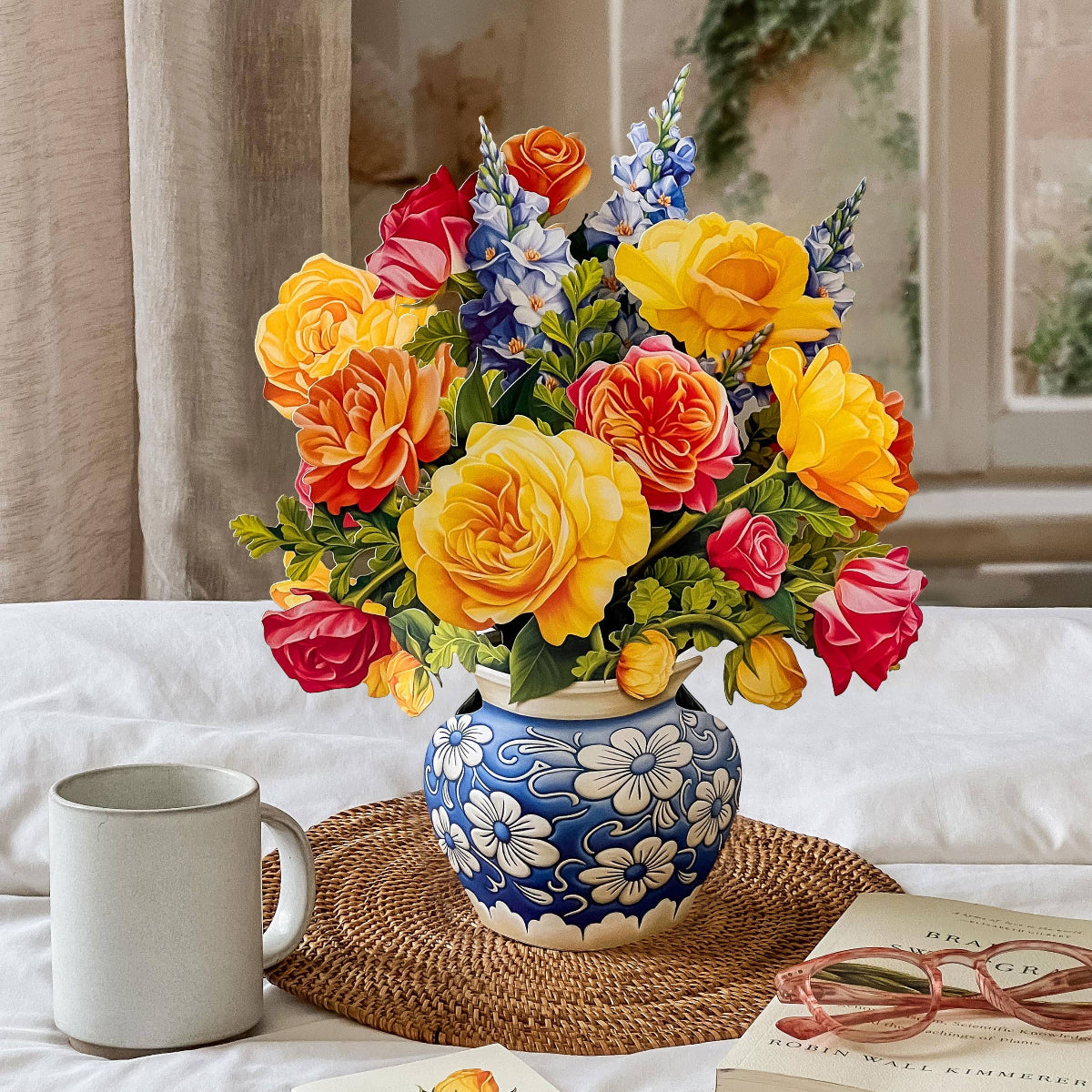 Colorful flower arrangement in a decorative vase on a table with a mug, pen, and book.