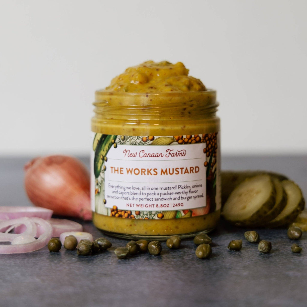 Jar of New Canaan Farms The Works Mustard with ingredients on a gray surface