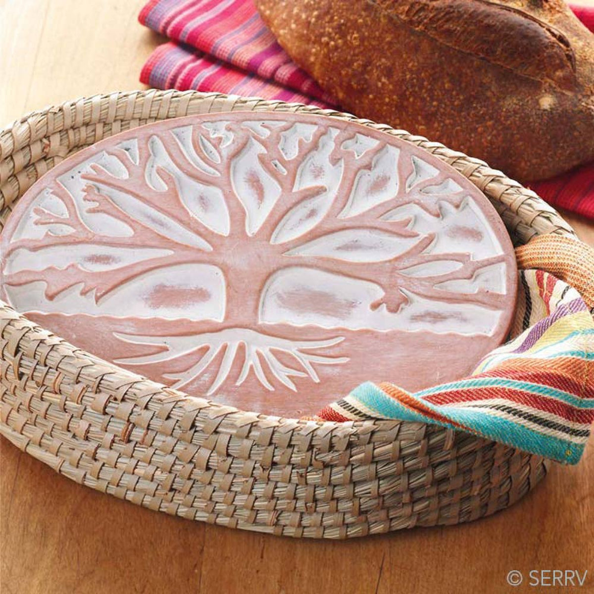 Decorative ceramic plate with tree design on a woven tray, surrounded by bread and colorful napkins.