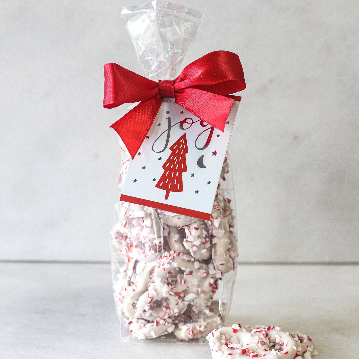 A clear plastic bag filled with white chocolate covered mini pretzels, decorated with red and green peppermint crunch, sealed with a red ribbon bow on top.