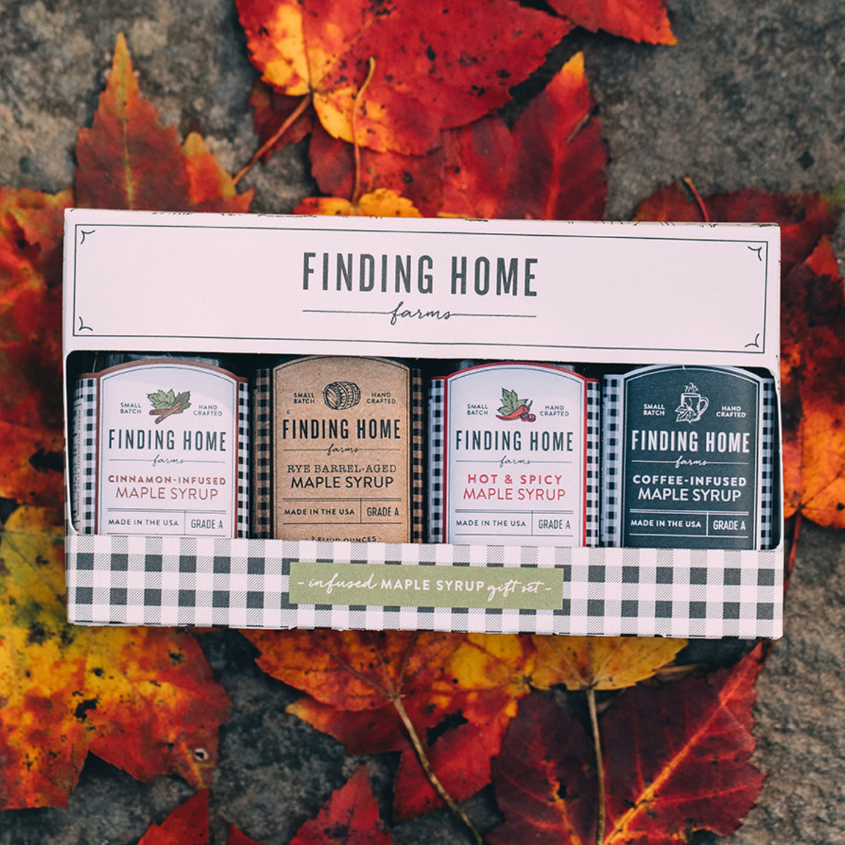 A boxed set of four Finding Home Farms maple syrup bottles displayed on a stone surface surrounded by red and orange autumn leaves. The set includes cinnamon-infused maple syrup, hot and spicy maple syrup, apple spice maple syrup, and coffee-infused maple syrup.